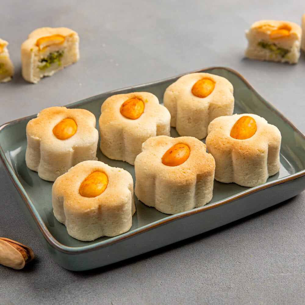 Tray filled with flower shaped badam biscuits each adorned with an almond piece. Around the tray are broken pieces of badam biscuits showing pista filling inside