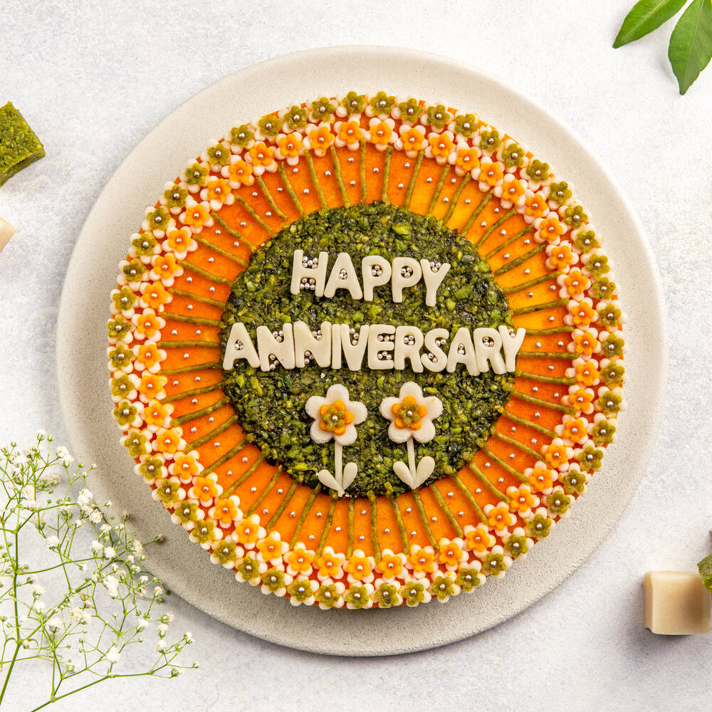 A cake made from badam and pista with intricate floral patterns and "Happy Anniversary" written in the center, perfect for a festive occasion.
