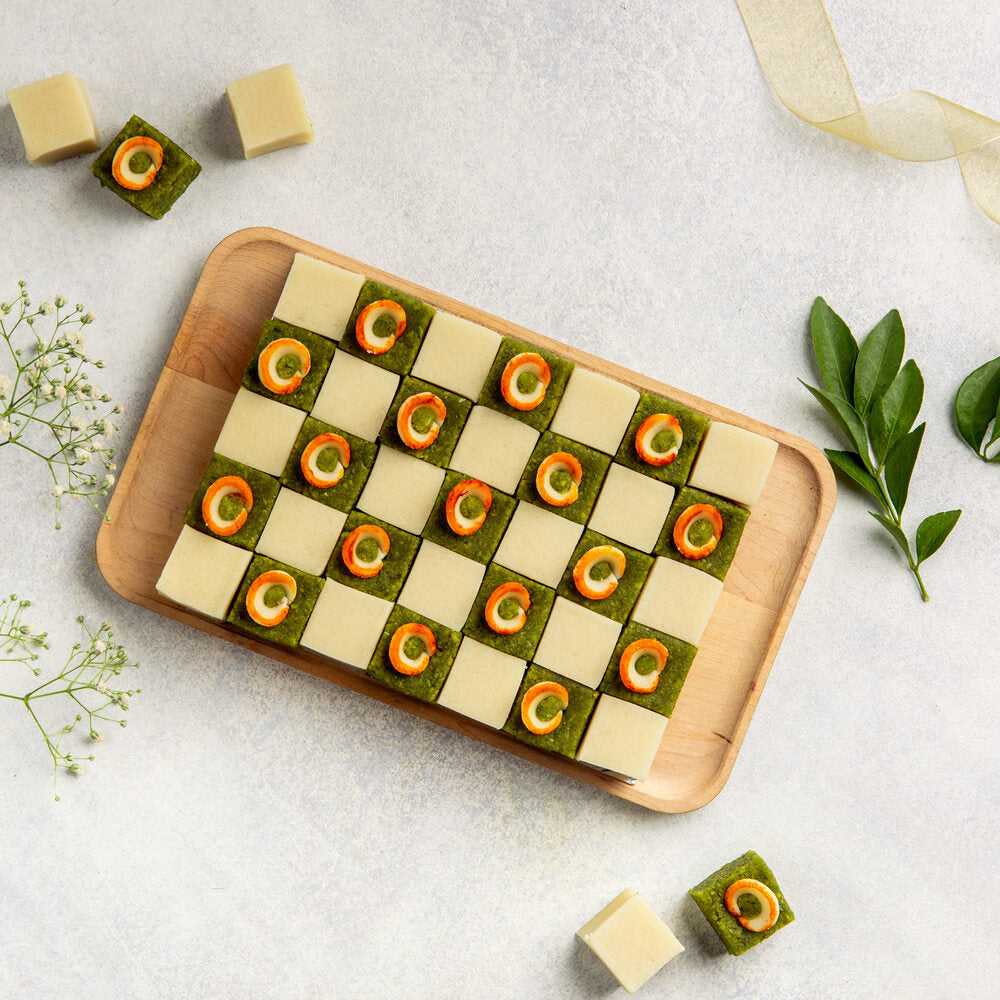 A check board display of alternating sqaure badam pista pieces. Each pista piece is topped with an orange and white tulip deoration