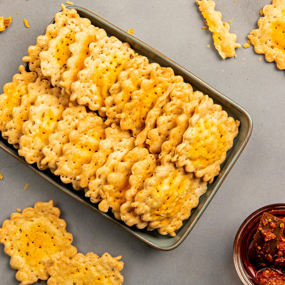 A tray of crispy, golden-baked achari mathri squares with scalloped edges. The crispy layers and savory pickled spices make them perfect for team time.