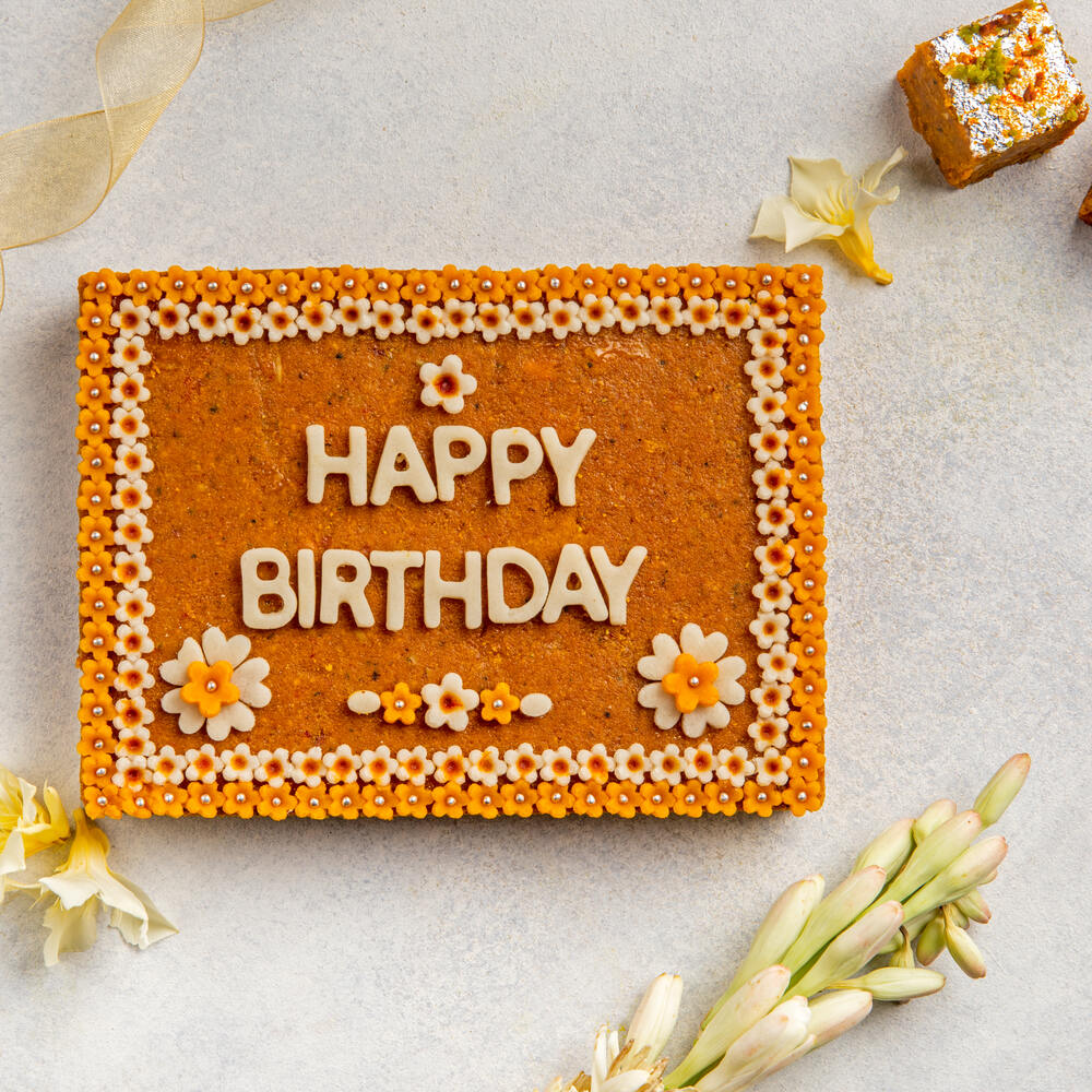 Beautifully crafted besan barfi in the shape of a cake with flower decoration and 'Happy Birthday' letters written on it