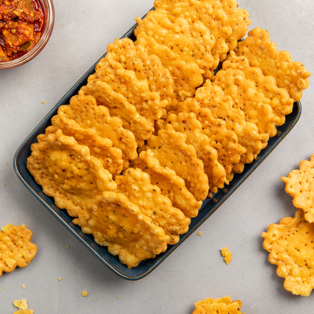 A tray of crispy, golden-fired achari mathri squares with scalloped edges. The crispy layers and savory pickled spices make them perfect for team time.